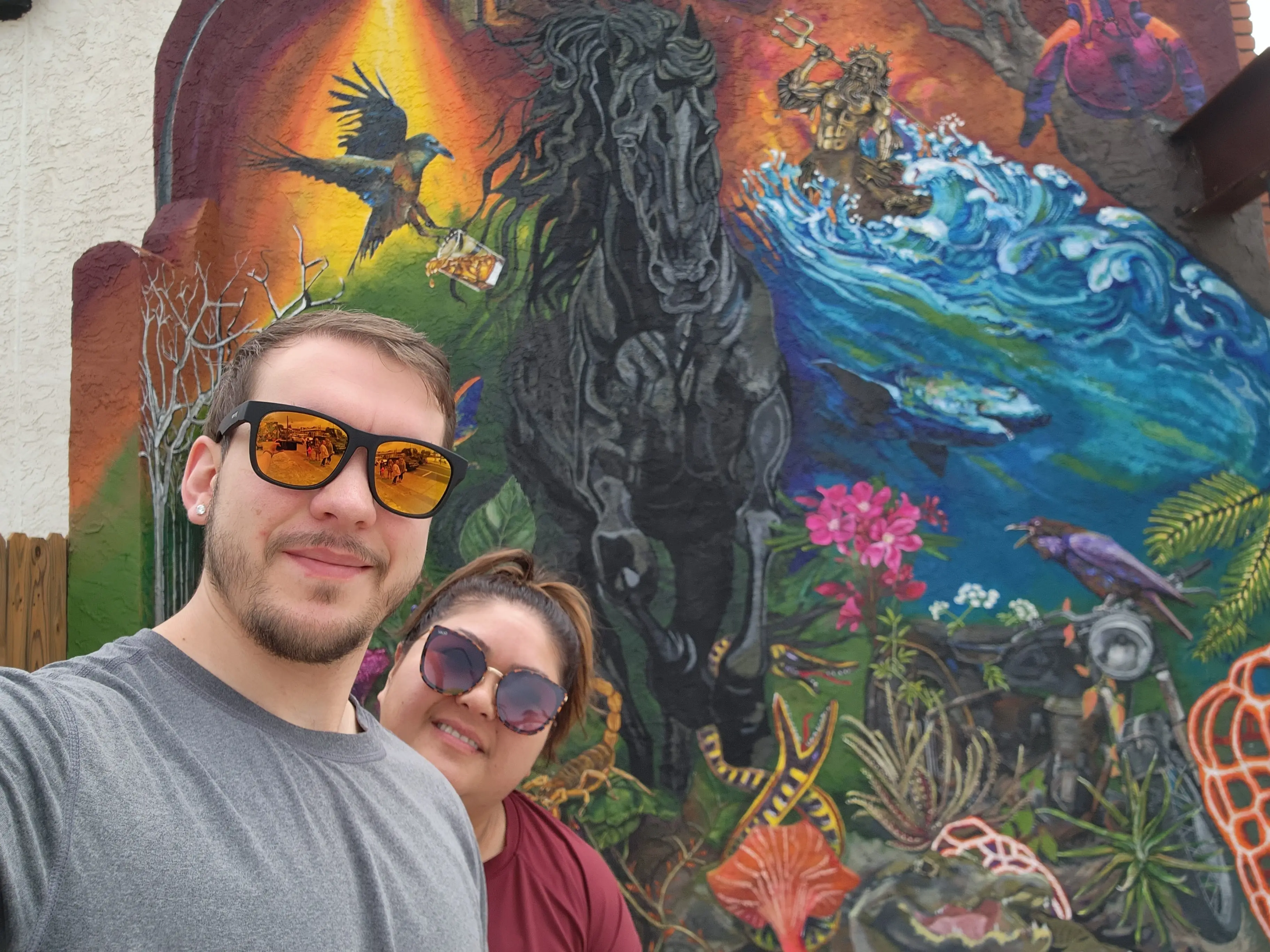 David Huson taking a selfie with a companion in front of a colorful mural of a black horse, ocean waves, and wildlife